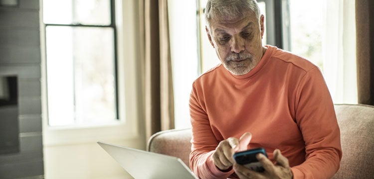 bild på man i orange tröja som sitter i soffan med mobil i handen och dator i knät